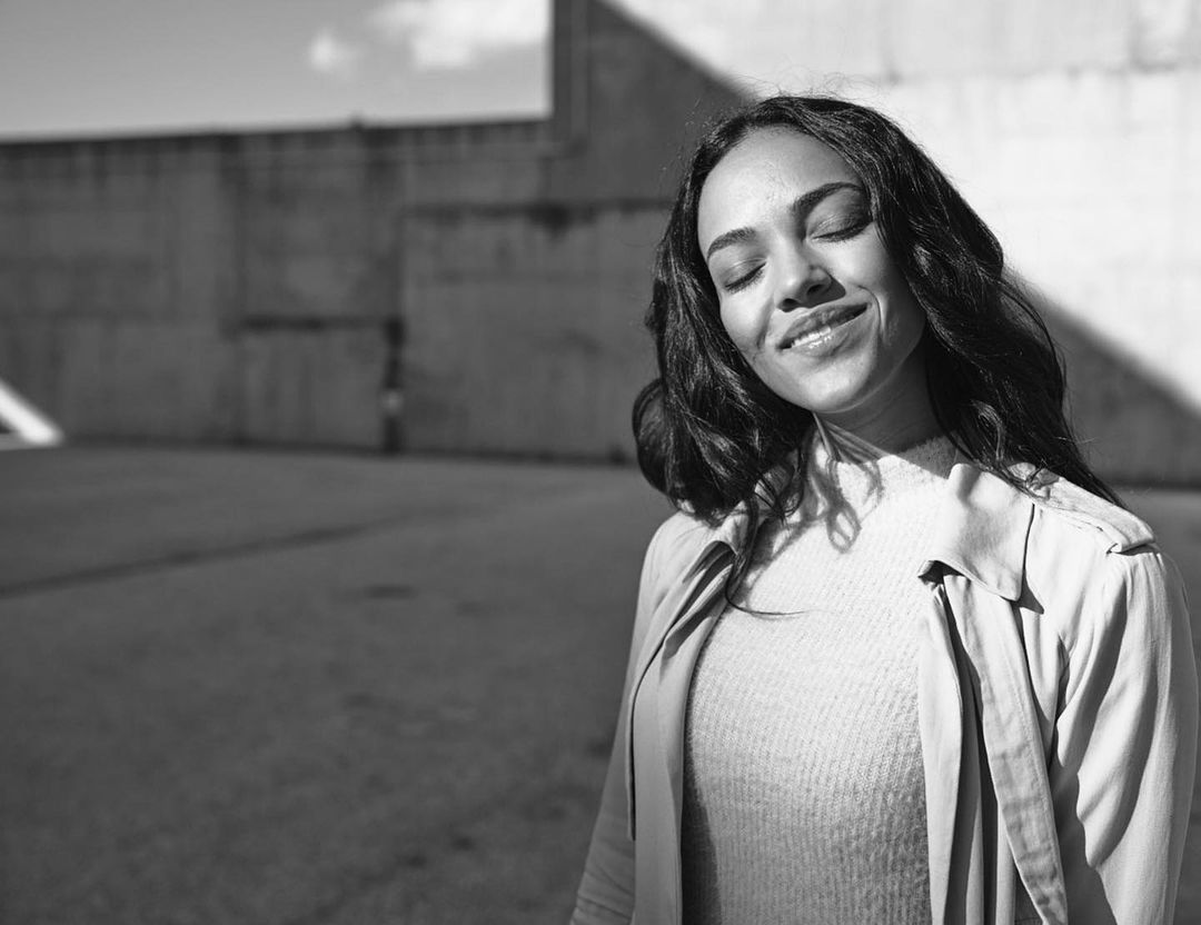 Black and white portrait of smiling adult woman with closed eyes wearing coat outside concrete building on sunny day.