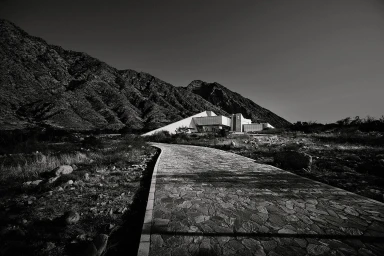Black and white photograph of a winding stone path leading toward a modern angular building nestled under rugged mountain slopes.