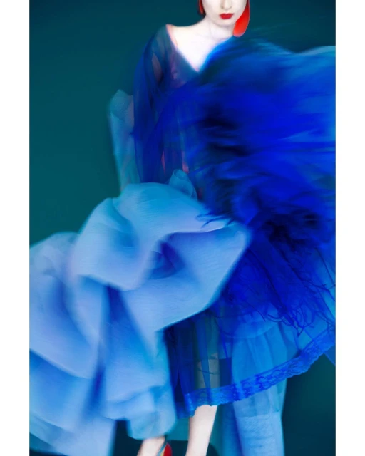 Adult female fashion model in motion wearing a blue flowing dress and red heels against a dark green studio background with motion blur.