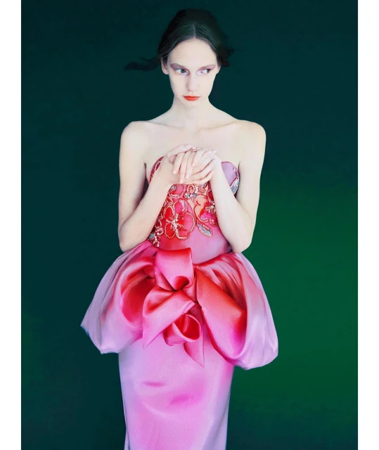 Elegant woman in pink gown with large bow poses against dark green backdrop, fashion editorial. Model wears strapless dress and red makeup.