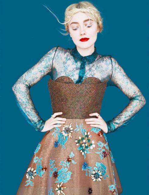 Woman posing in an elaborate dress with lace sleeves and floral accents against a teal background in a fashion editorial.