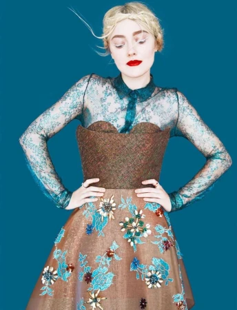Woman posing in an elaborate dress with lace sleeves and floral accents against a teal background in a fashion editorial.