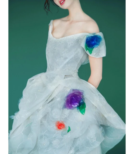 A fair-skinned adult woman is shown in a ruffled white dress with flower details on a green background in a portrait shot.