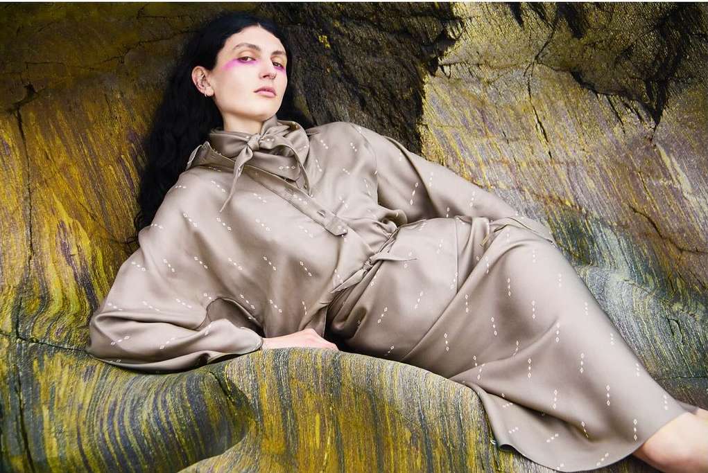 Woman in beige patterned dress poses against a textured rock formation, illuminated by natural light, showcasing fashion and style.