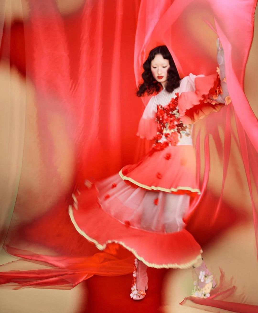 A woman in a flowing coral gown poses among red and pink sheer fabric. She has a fair complexion with dark hair and red lipstick.