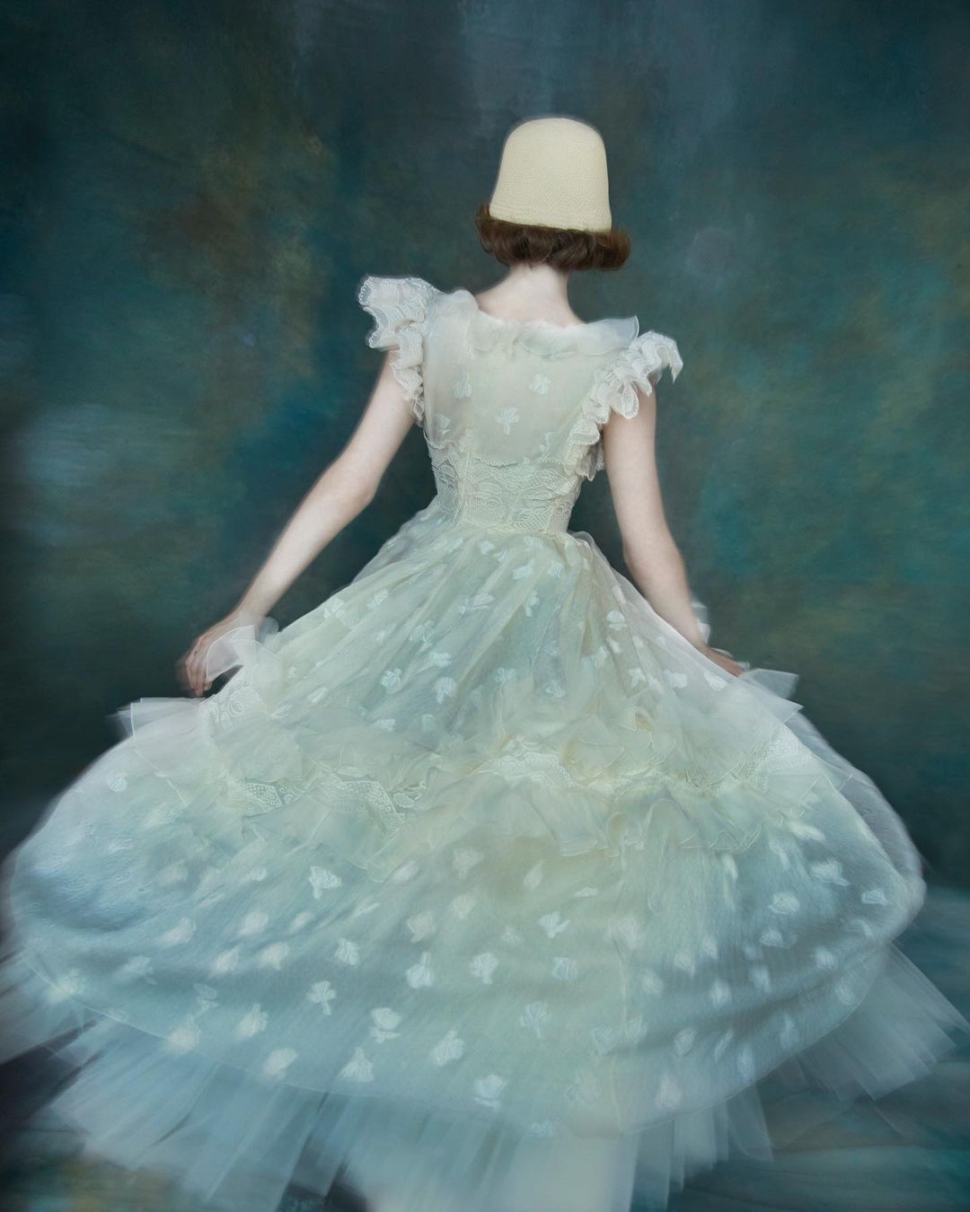 Back view of a person in a white floral dress and a unique hat posing in front of a mottled blue and brown background.