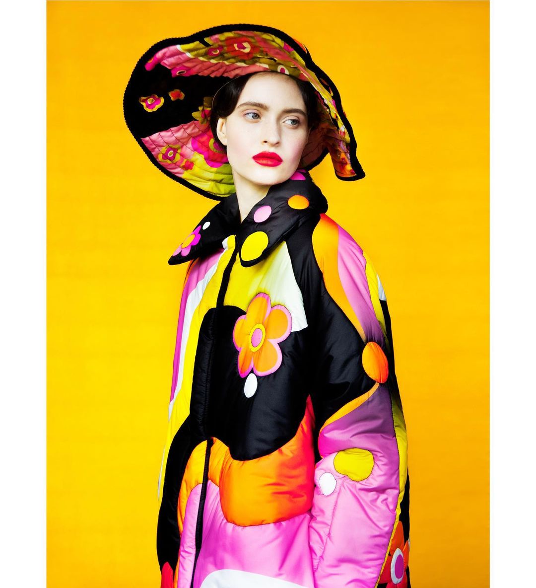 Woman wearing a bright floral hat and a flower-power patterned puffer jacket against a solid yellow backdrop in a studio setting.