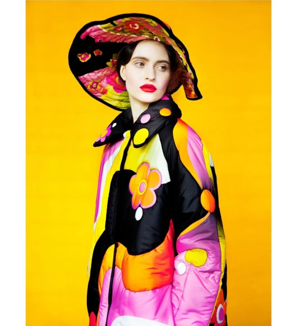 Woman wearing a bright floral hat and a flower-power patterned puffer jacket against a solid yellow backdrop in a studio setting.