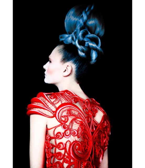 Avant-garde fashion portrait of a woman with blue braided hair, wearing a red decorative dress against a dark black studio backdrop.