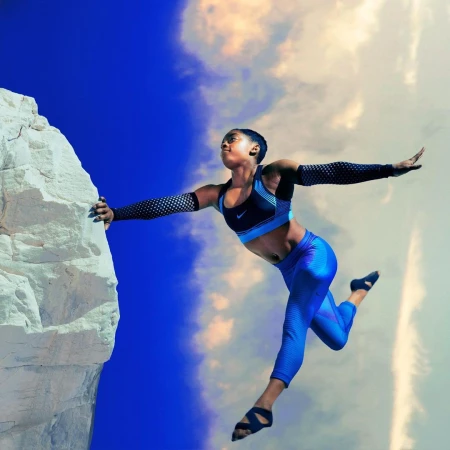 Athletic African-American woman leaps from rock cliff against a vibrant blue sky with wispy clouds wearing blue athletic wear, sleeves, and open toe sandals.