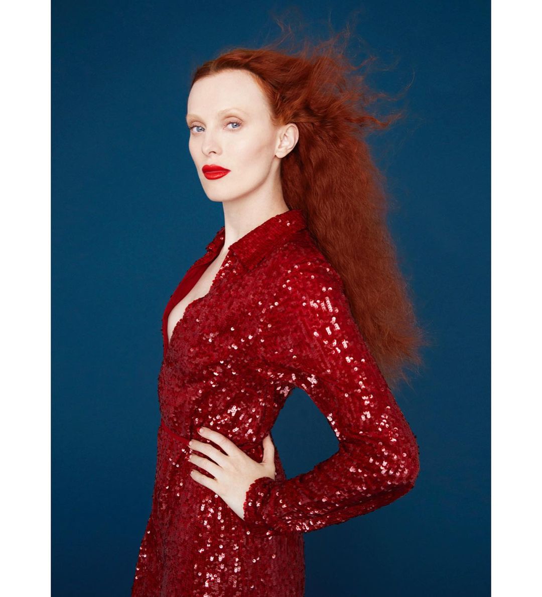 Side profile of a red-haired adult woman in a red sequined dress against a dark blue backdrop in a studio portrait.