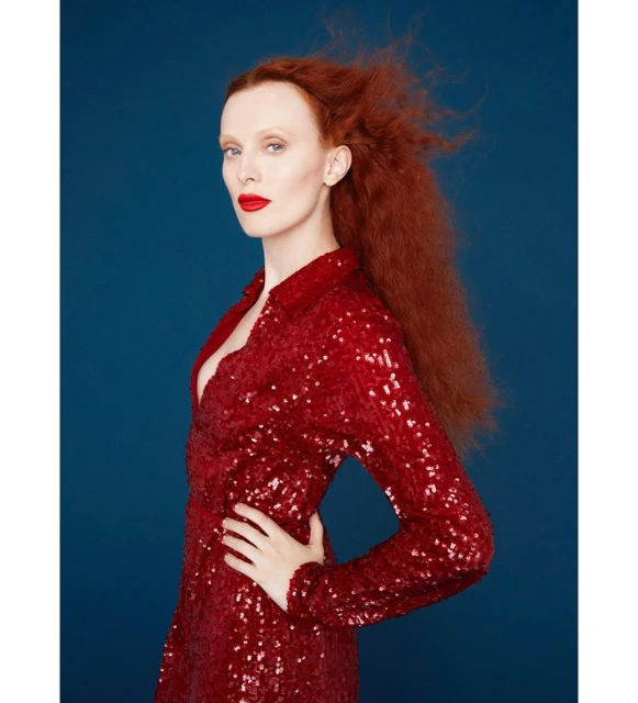 Side profile of a red-haired adult woman in a red sequined dress against a dark blue backdrop in a studio portrait.