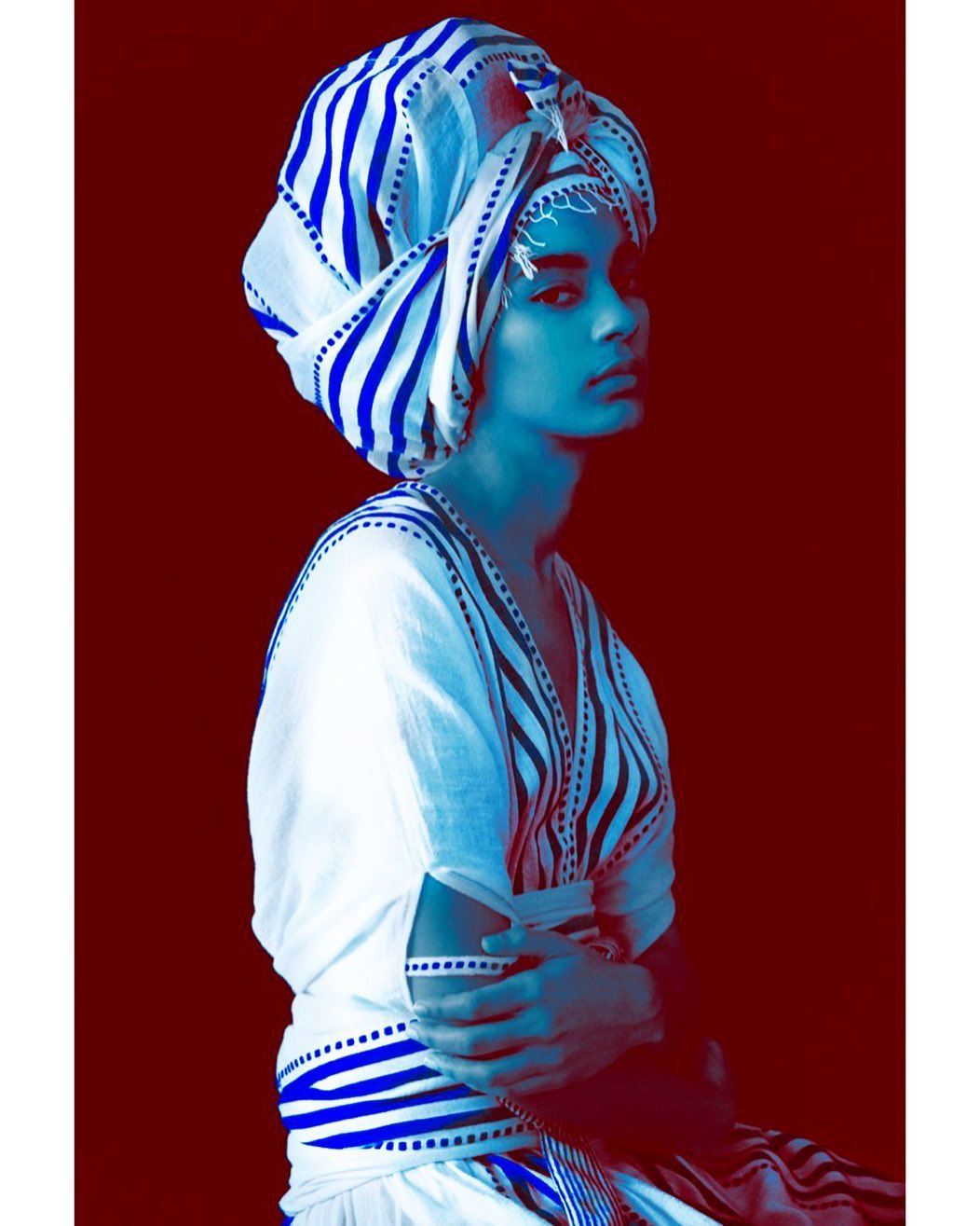 A young adult in traditional headwear poses in a studio shot against a dark red background, showcasing blue and white striped fabric.