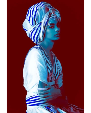 A young adult in traditional headwear poses in a studio shot against a dark red background, showcasing blue and white striped fabric.