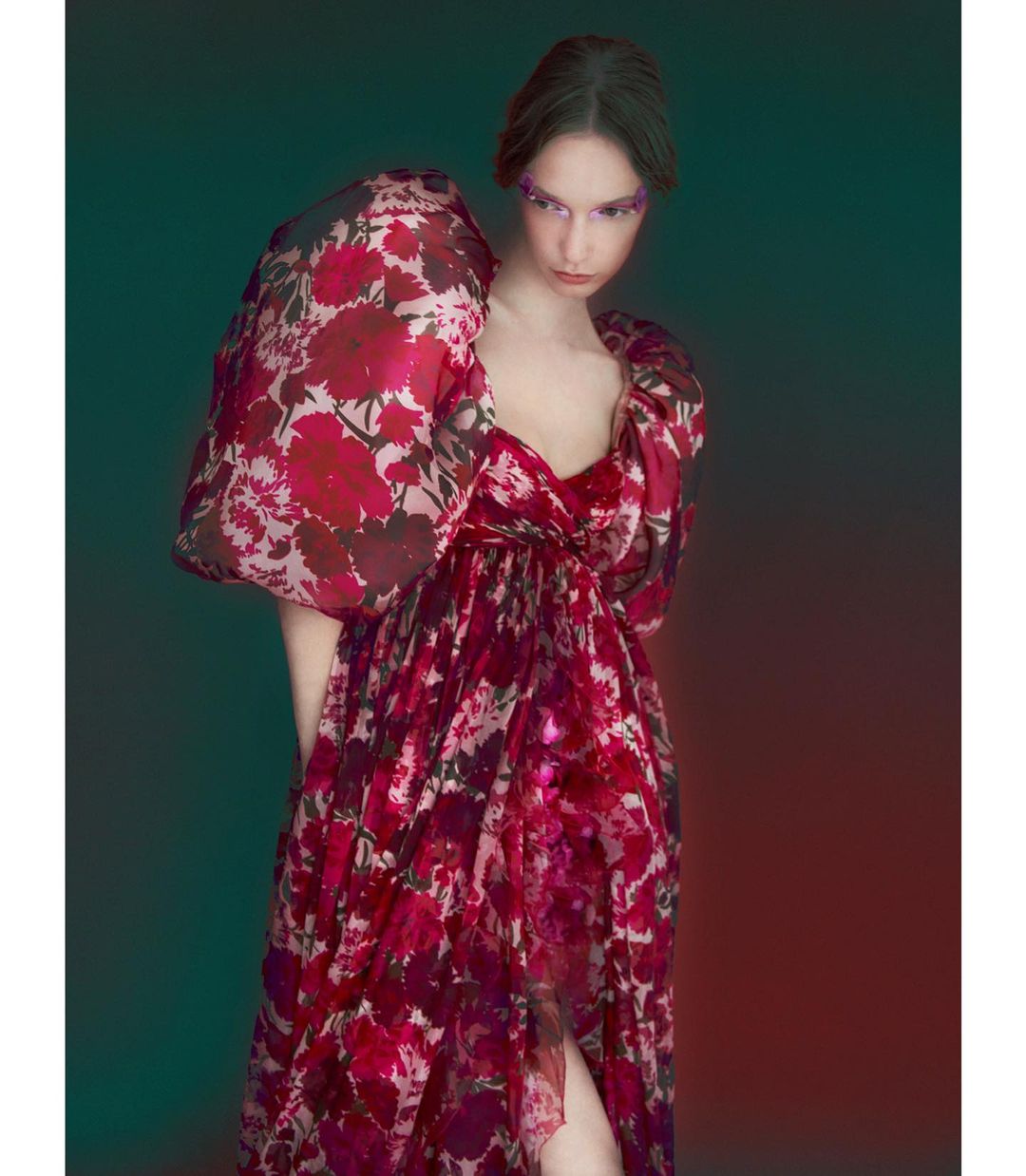 Woman in elegant red floral gown with dramatic puffy sleeves and glitter eye makeup poses against a gradient background in a studio portrait.