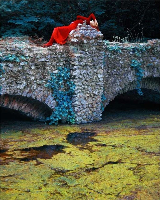 Woman in red dress lies on a stone bridge over a green, mossy body of water, surrounded by dark green trees, in an outdoor setting.