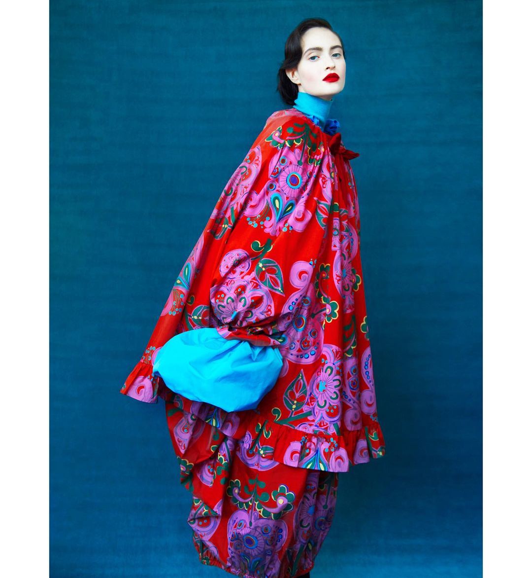 Woman in vibrant, patterned dress and bright blue accessory posing against a solid teal backdrop in a fashion editorial style photograph.