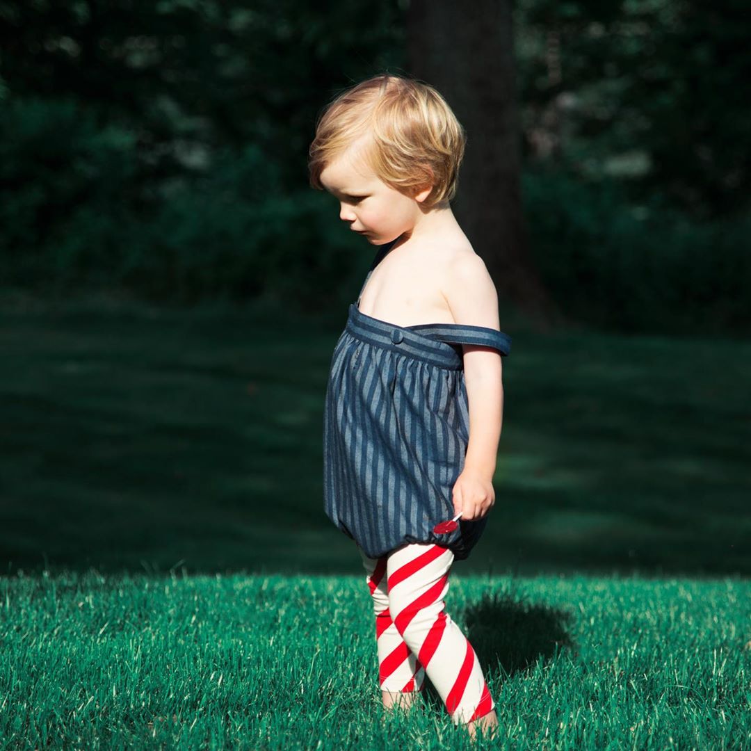 Young child with blonde hair stands outside in blue striped top and red striped leggings in a grassy outdoor setting looking down.