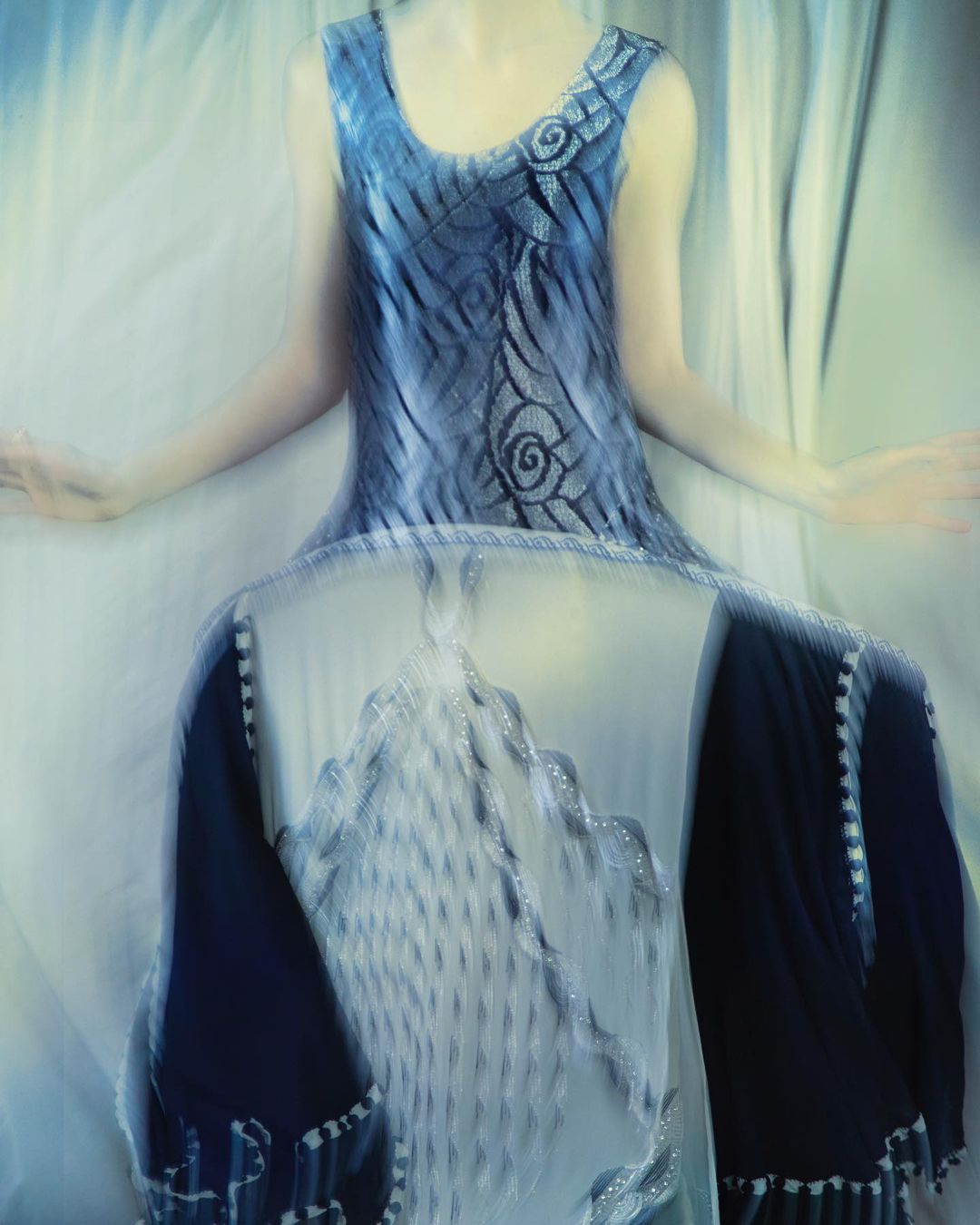 Abstract fashion shot featuring a blurred female model wearing a sleeveless blue dress with decorative swirls against a soft white background.