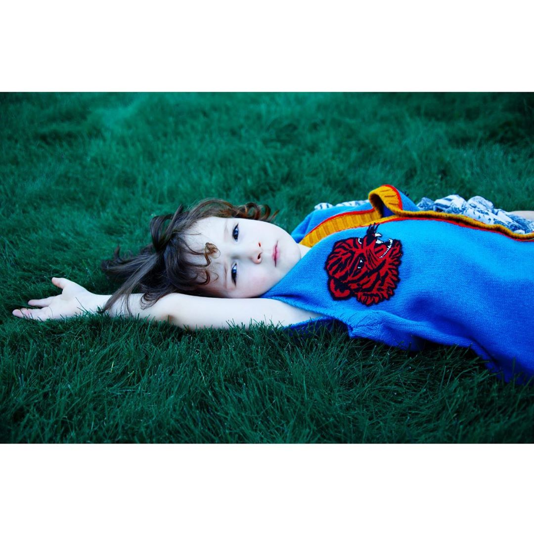 Young child lies on a vibrant green lawn, gazing peacefully at the camera in a close-up shot on a bright day, with a blue patterned shirt.