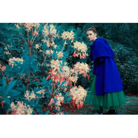 A young woman in a blue coat and green skirt poses in a lush garden with white and coral flowers on bushes, creating a fashion editorial.