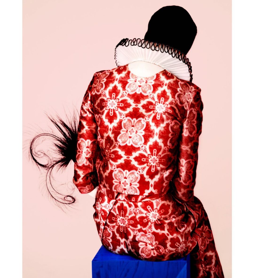Back view of a person in an ornate red and white patterned jacket, white ruff collar, and black cap, sitting on a blue stool against pink.