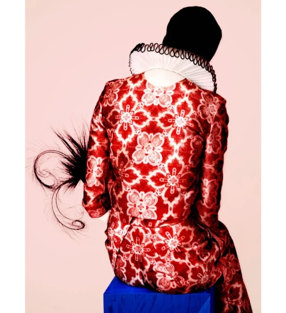 Back view of a person in an ornate red and white patterned jacket, white ruff collar, and black cap, sitting on a blue stool against pink.
