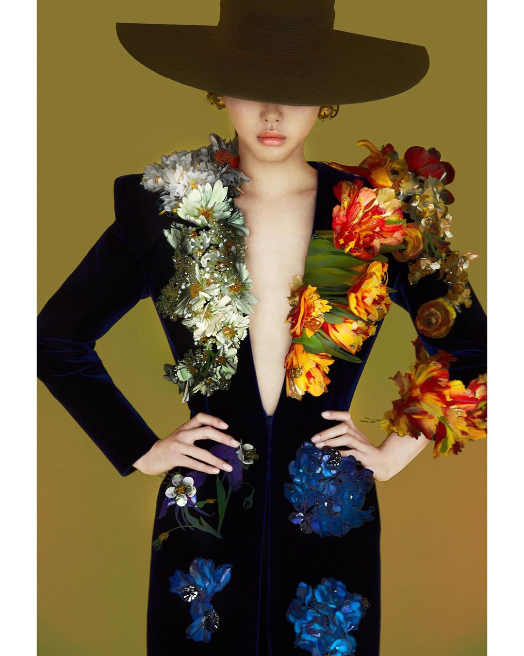 Woman in dark blue velvet dress with floral embellishments and wide-brimmed hat stands against a muted yellow studio backdrop.