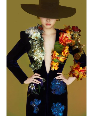 Model wearing a dark velvet jacket adorned with floral embellishments and a wide-brimmed black hat against a solid background