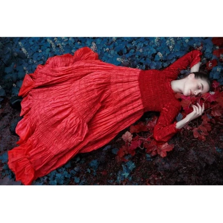 Woman in a red dress lies on the ground surrounded by colorful leaves in a close-up, high-angle shot showing her in an outdoor setting.