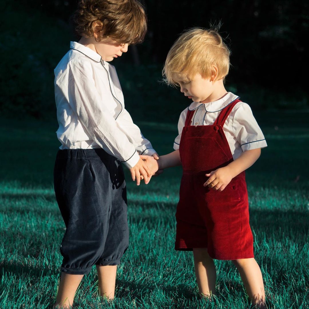 Two young children stand on grassy field, holding hands. One wears white top and blue pants, other wears white top and red overalls.