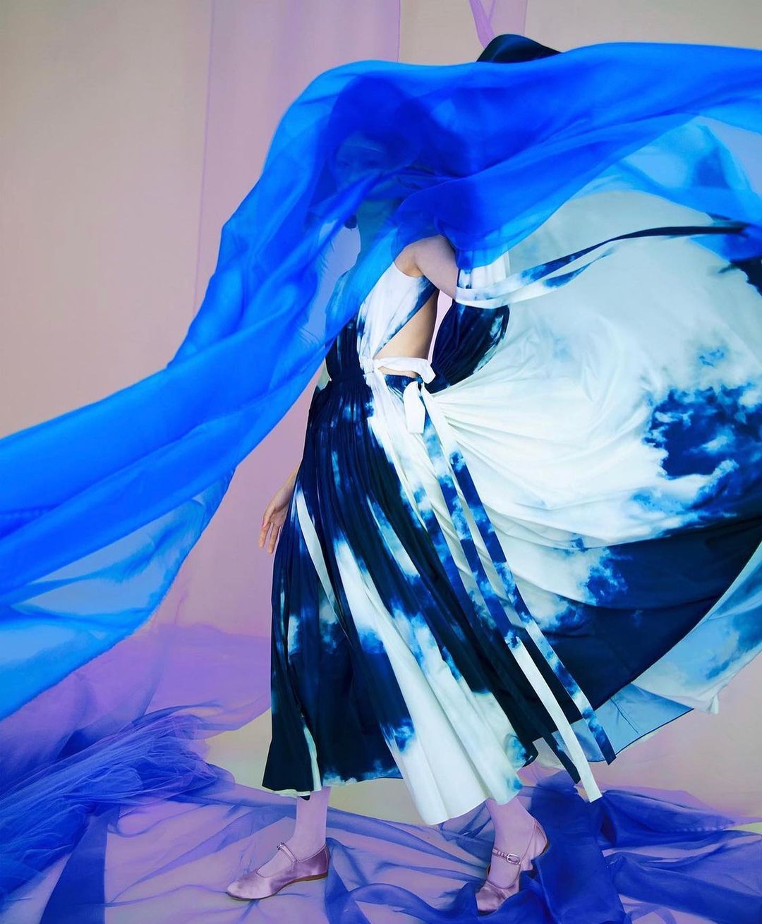 Fashion editorial featuring a person in a flowing tie-dye dress and a blue sheer fabric against a neutral background in a studio setting.