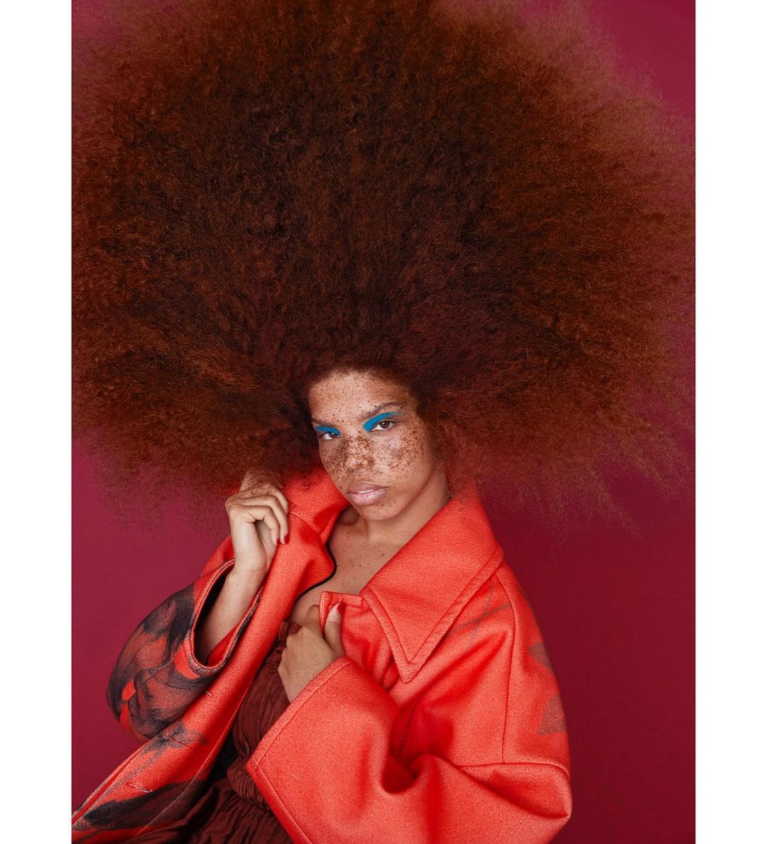 A striking portrait of a freckled woman with a large auburn afro, wearing bright blue eyeshadow and a red-orange coat against a red backdrop.