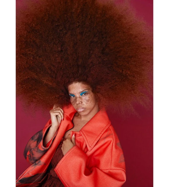 A striking portrait of a freckled woman with a large auburn afro, wearing bright blue eyeshadow and a red-orange coat against a red backdrop.