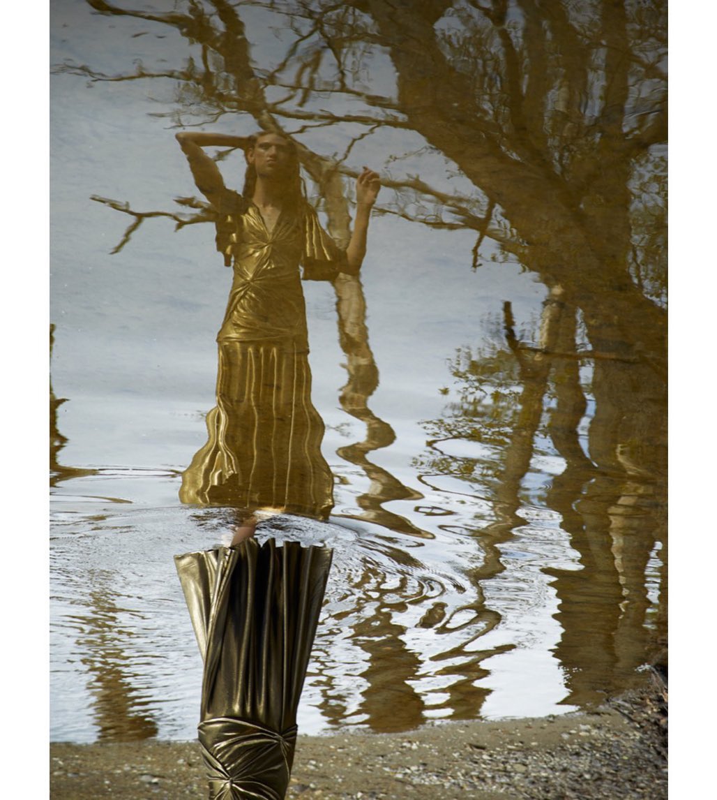 Fashion shot of a woman in a long, golden gown, reflected in rippling water with tree reflections creating an ethereal, artistic scene.