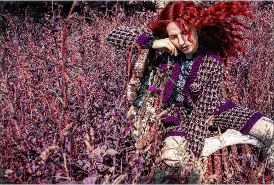A fair-skinned adult woman with curly red hair in a houndstooth knit outfit is seated on a wooden bench surrounded by thick weeds.