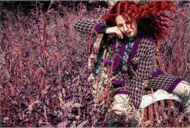 A fair-skinned adult woman with curly red hair in a houndstooth knit outfit is seated on a wooden bench surrounded by thick weeds.