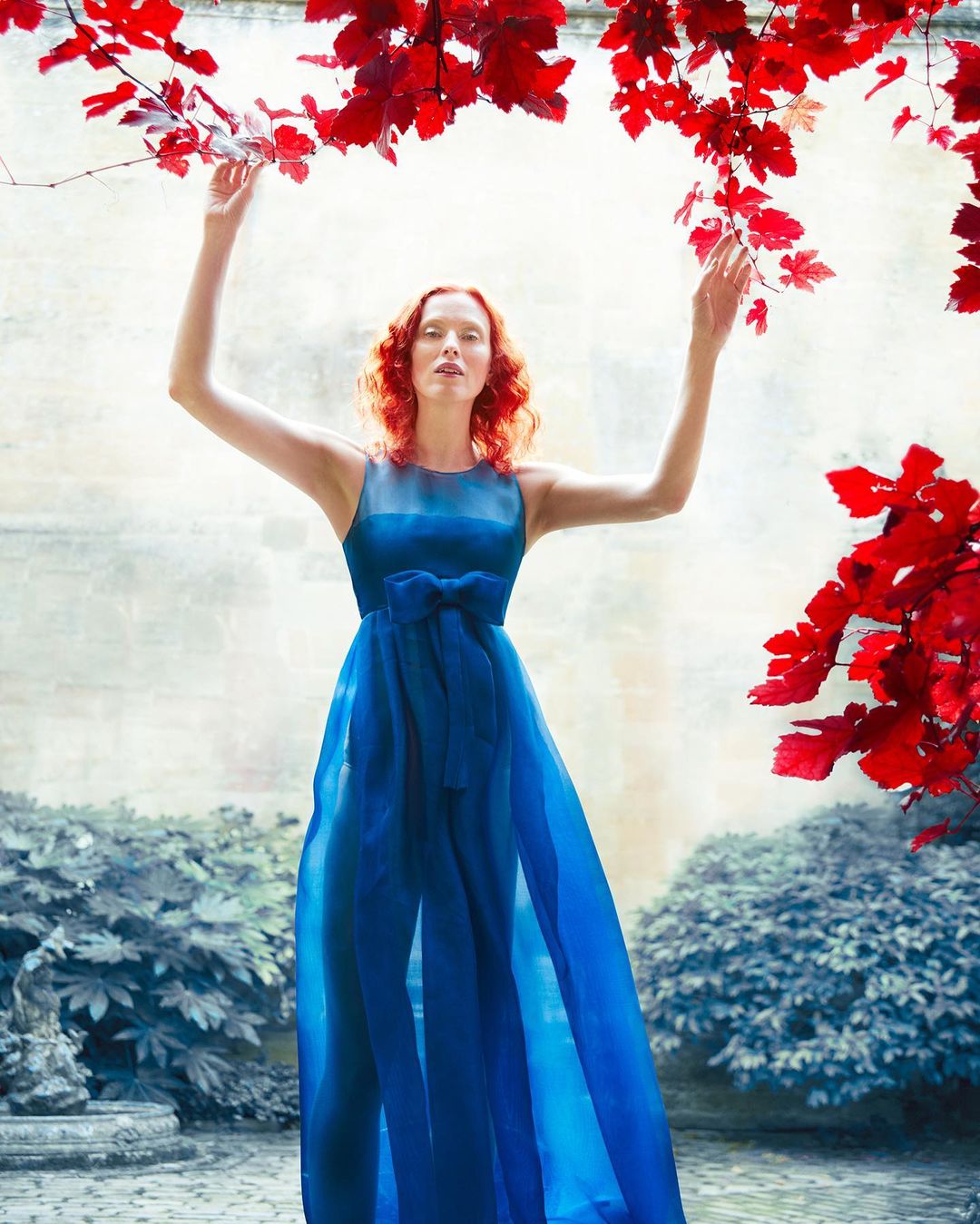 Woman in a flowing blue dress stands in a courtyard with red foliage above. Fashion editorial with a surreal and artistic aesthetic.