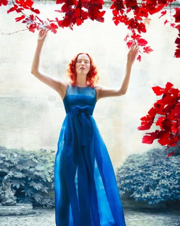 Woman in a flowing blue dress stands in a courtyard with red foliage above. Fashion editorial with a surreal and artistic aesthetic.