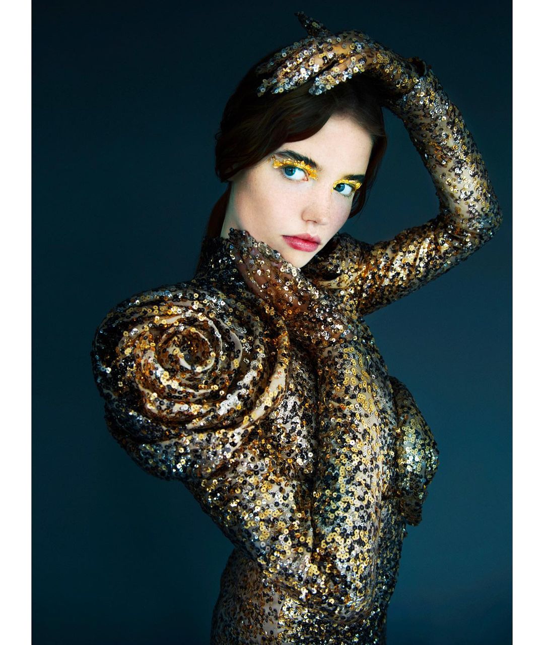 A woman in a sparkling gold dress poses in front of a dark gray background, wearing bright yellow eyeshadow and placing her hands up by her head.