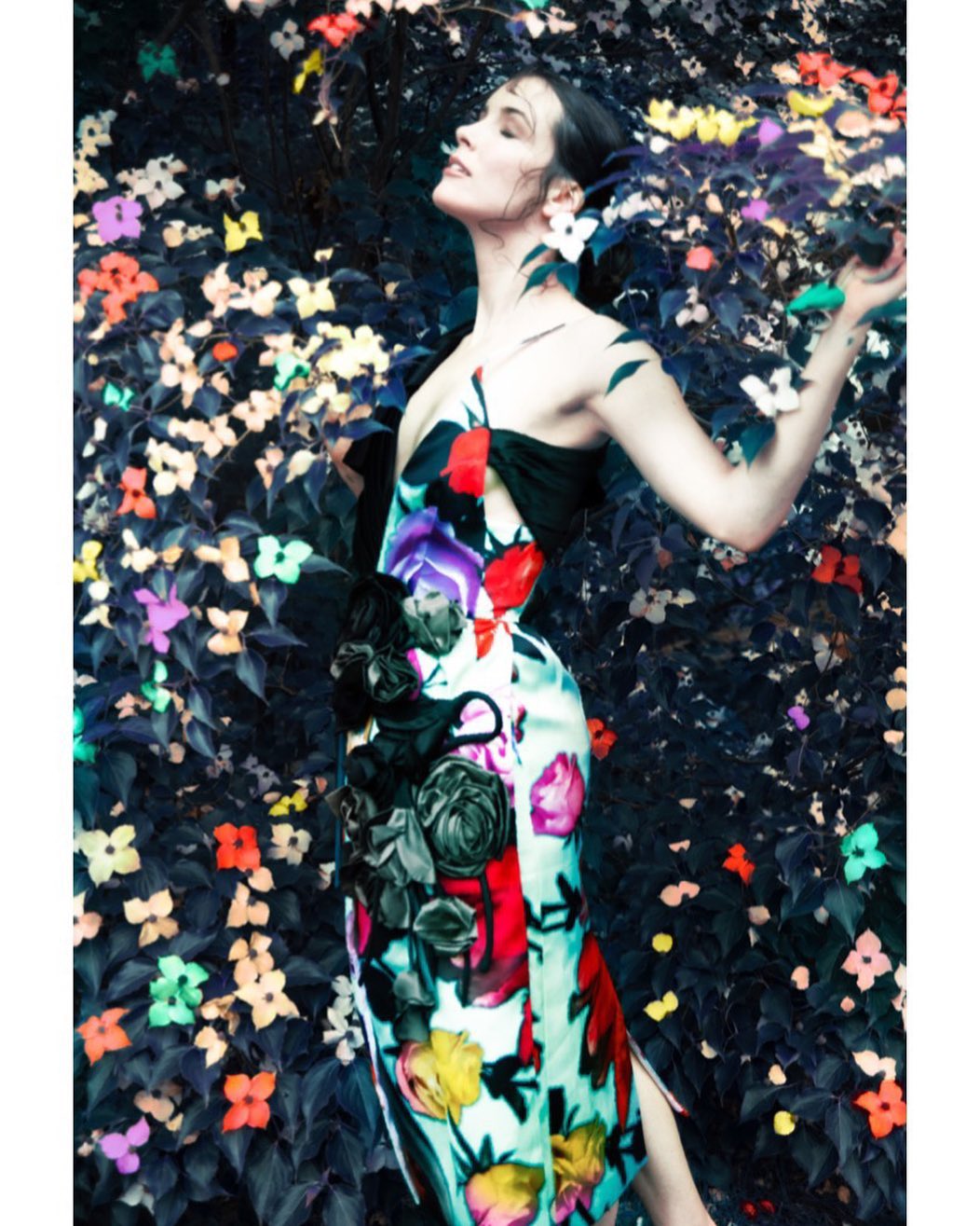 Woman in floral gown in garden. Model in outdoor fashion shoot surrounded by flowers and leaves, eyes closed, natural light.