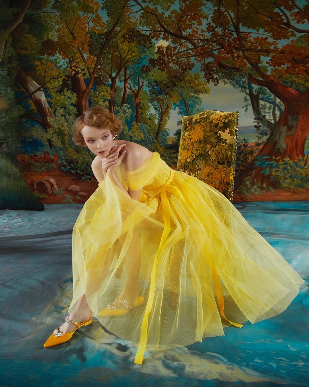 Woman in yellow tulle dress sits on patterned chair in front of an artful backdrop depicting a forest and stream.