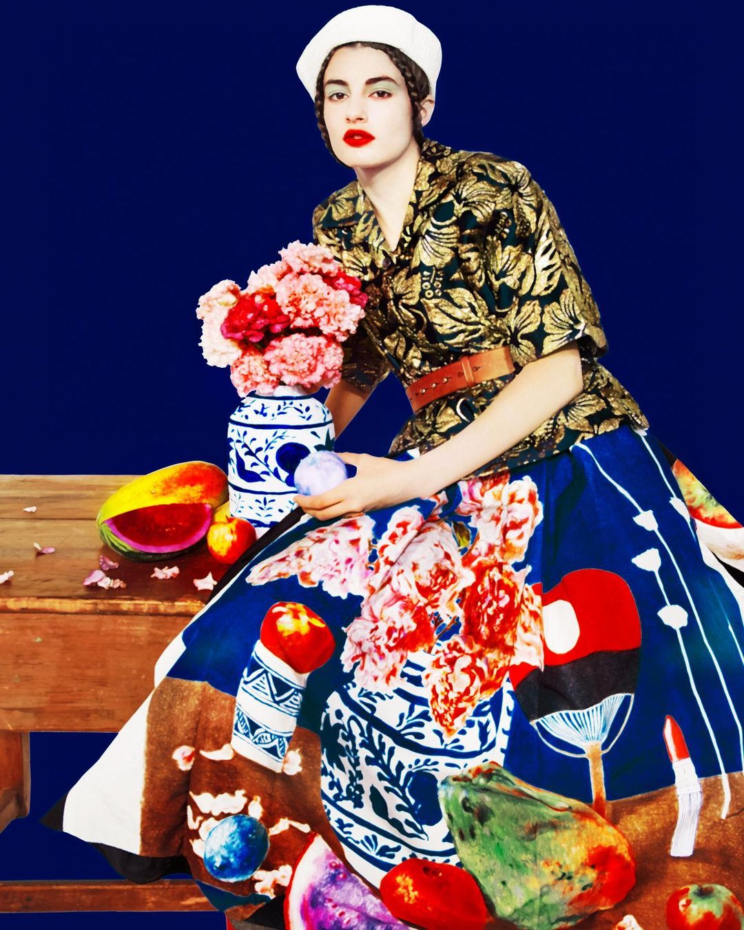 Woman in floral patterned dress and beret poses with flower-filled vase in a fashion editorial studio shot against deep blue backdrop.