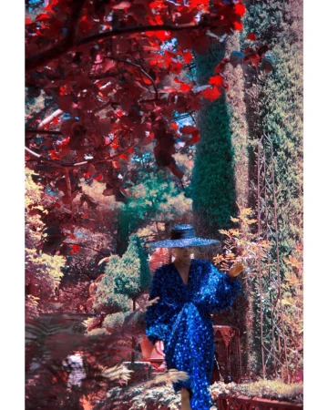 Woman in a sparkling blue dress and hat sits on a ledge surrounded by lush greenery and red foliage in an outdoor garden setting.