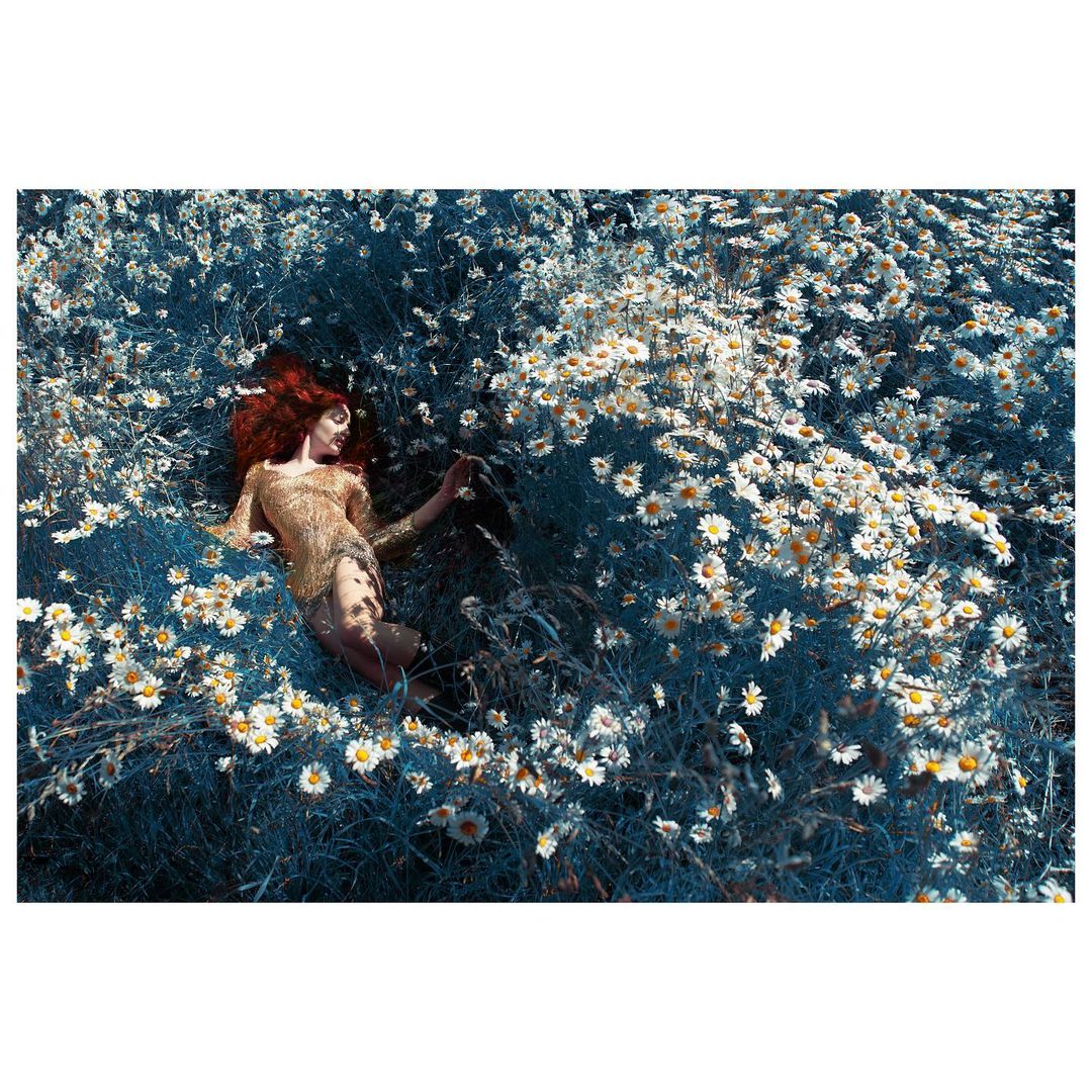 Redhead woman in a gold dress lies in a field of white daisies, bathed in soft light, creating a dreamy and ethereal atmosphere.