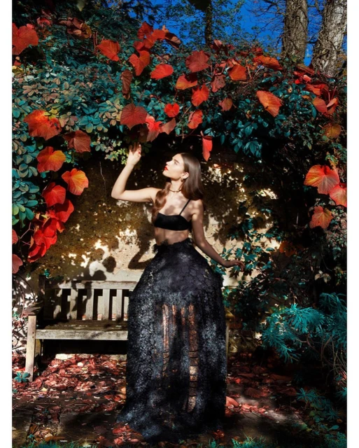Woman poses in a black lace gown, touching red autumn leaves, in an outdoor nature setting, beside an old wooden bench.