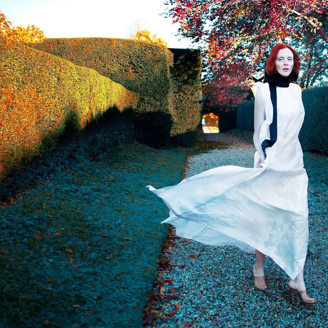 Woman in long white dress walks along a path in a formal garden with tall hedges, autumn colors, and textured surface.