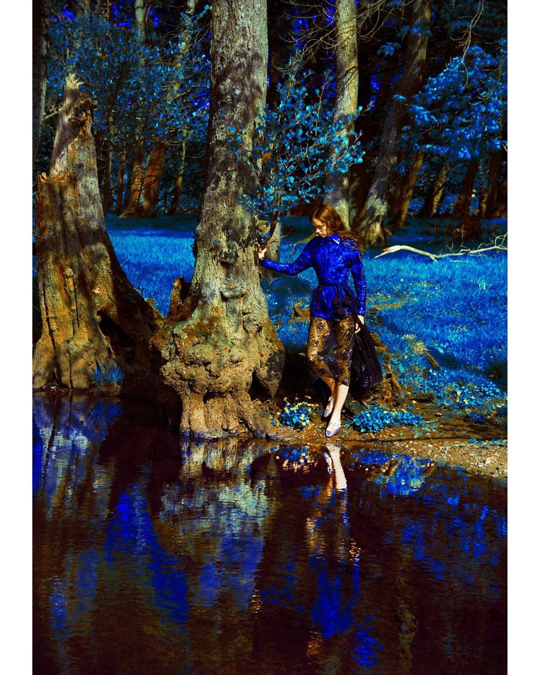 Woman in blue jacket poses by reflective water in a dreamy forest scene with tall trees and a vibrant blue color palette.