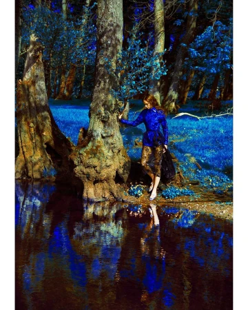 Woman in blue jacket poses by reflective water in a dreamy forest scene with tall trees and a vibrant blue color palette.