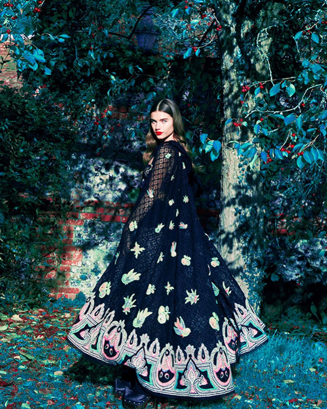 An adult female model poses outdoors wearing a black cape with floral embellishments, standing in a lush garden with a brick wall.